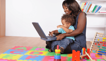 woman holding toddler on child's mat and working on computer