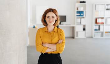 redheaded petite woman in yellow blouse stands, smiling, with arms crossed in office