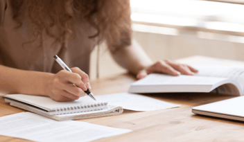 woman with brown curly hair, writing on notepad, with book open